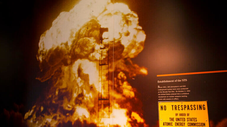 A large mushroom cloud rises from a nuclear explosion, with a NO TRESPASSING sign by order of the U.S. Atomic Energy Commission visible in the foreground, warning against unauthorized entry.