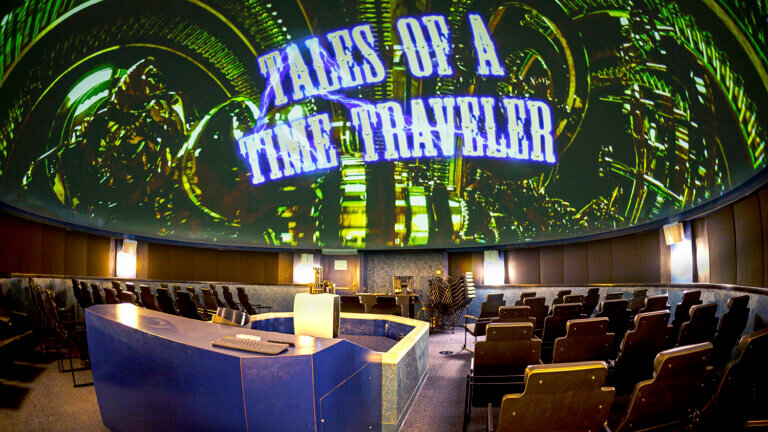 A planetarium theater with empty seats faces a large domed screen displaying the title Tales of a Time Traveler in bold letters over a sci-fi background. A blue control desk sits at the front of the room.