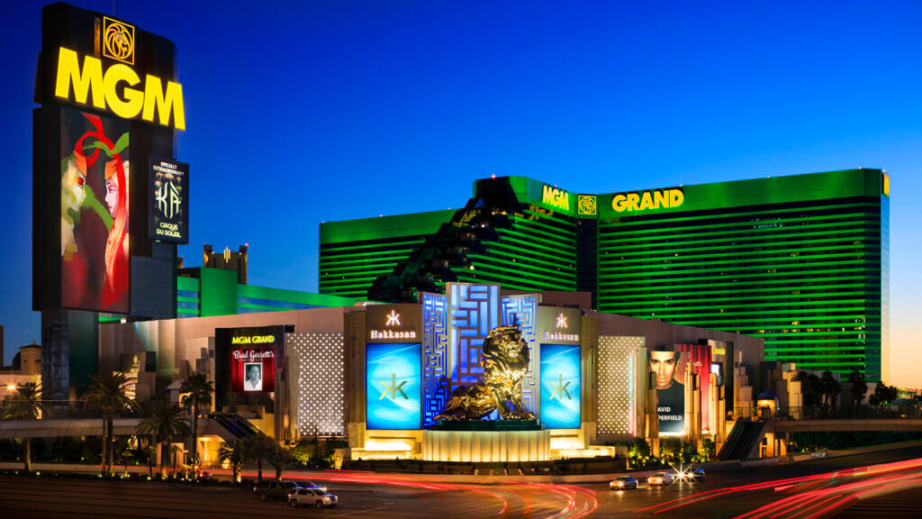 The MGM Grand hotel and casino in Las Vegas is brightly lit at night, featuring a large golden lion statue in front, digital billboards, and green lights highlighting the building’s name.