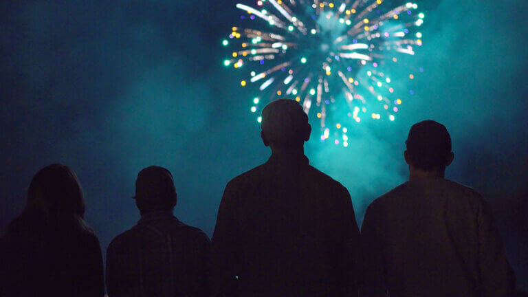 Four people in silhouette watch bright, colorful fireworks exploding in the night sky, surrounded by smoke and illuminated by the glow of the display.