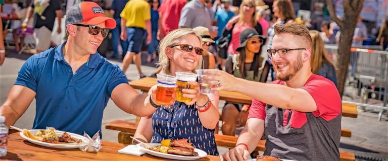 Three people toast with drinks at an outdoor festival, sitting at a wooden table with food and smiling.