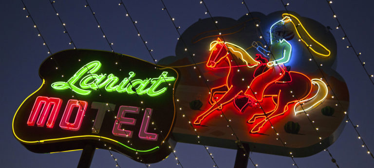 A neon sign at night reads Lariat Motel in green and pink, with a colorful neon image of a cowgirl riding a horse, outlined by small string lights above.