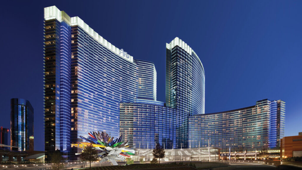 Modern, glass-clad high-rise buildings illuminated at night with a colorful abstract sculpture in the foreground, set against a clear dark blue sky.