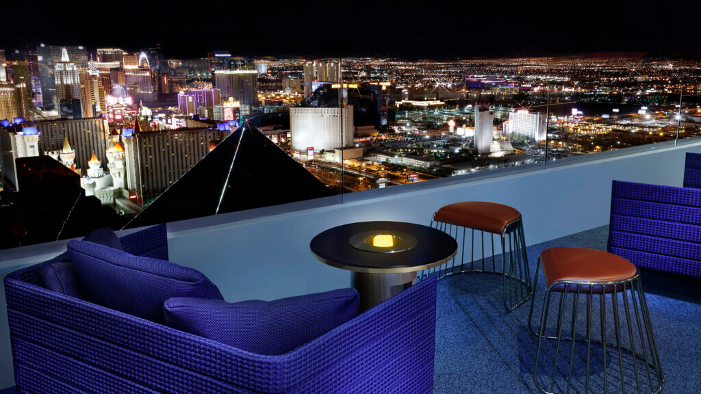A rooftop lounge with modern purple chairs and stools overlooks a dazzling, illuminated Las Vegas cityscape at night through clear glass panels.