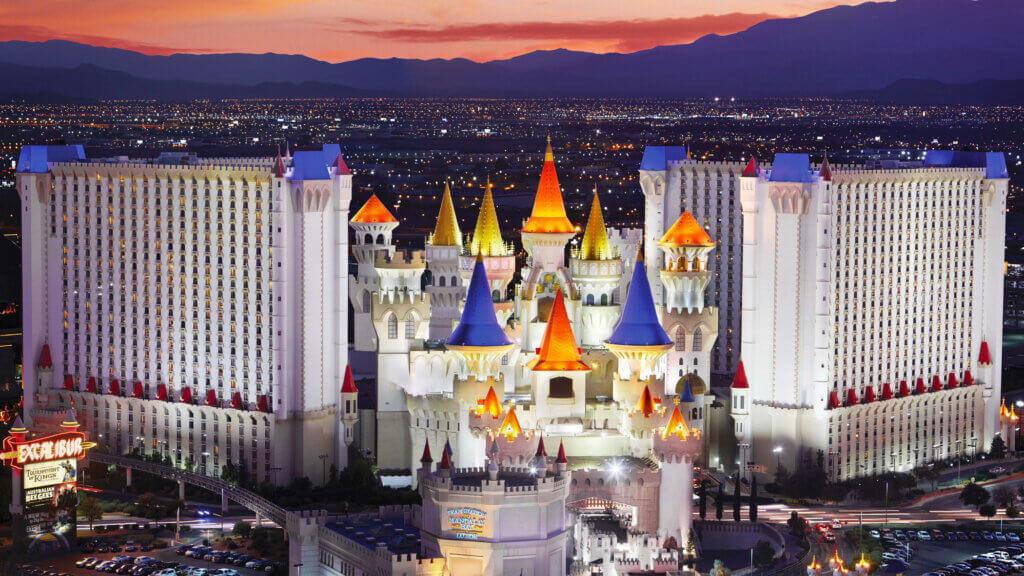 A brightly lit castle-themed hotel with colorful turrets stands against a sunset sky and city lights in Las Vegas, with mountains in the background.