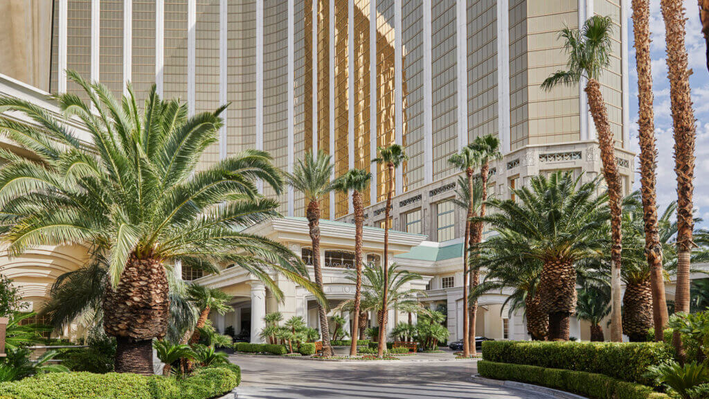 Tall, modern hotel buildings with gold-tinted windows surrounded by palm trees and lush greenery, with a curved driveway in the foreground under a partly cloudy sky.