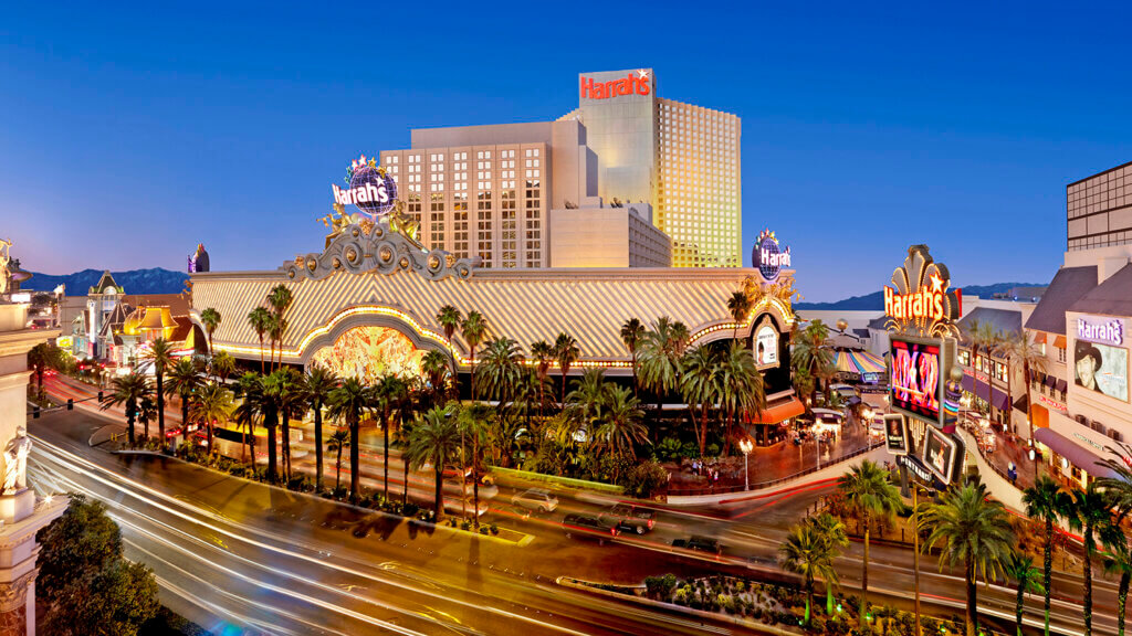 A vibrant, evening view of Harrah’s Las Vegas hotel and casino, with bright lights, palm trees, and colorful signage along a busy street on the Las Vegas Strip.