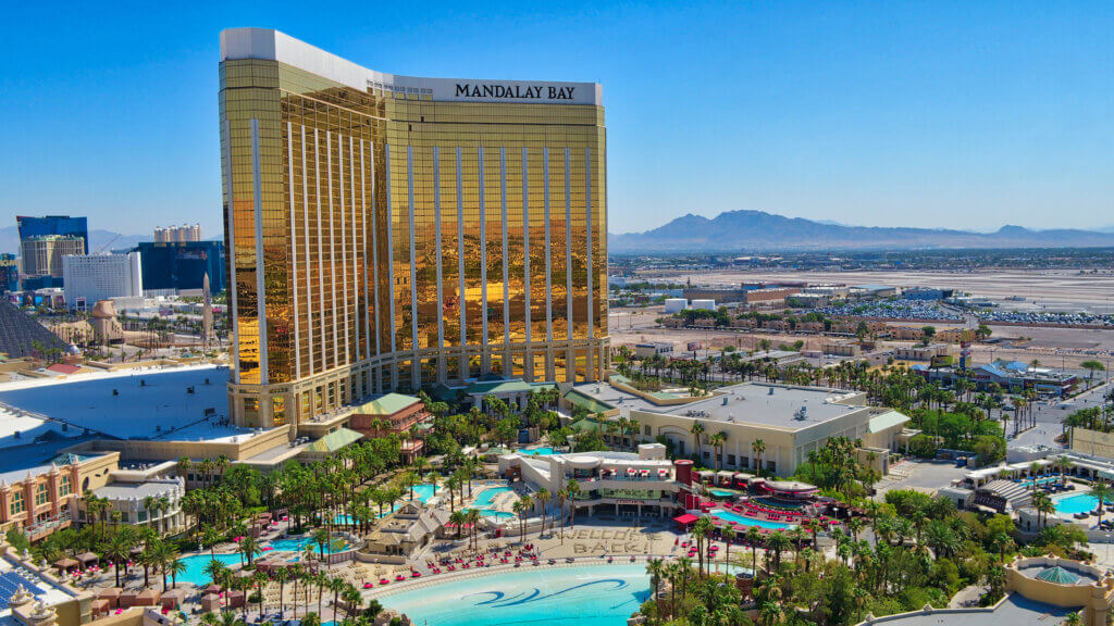 Aerial view of the Mandalay Bay hotel and casino in Las Vegas, featuring its tall gold glass building, outdoor pools, palm trees, surrounding resort grounds, and distant mountains under a clear blue sky.