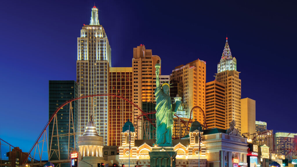 Colorful night view of the New York-New York Hotel & Casino in Las Vegas, featuring replicas of New York skyscrapers, the Statue of Liberty, and a looping red roller coaster.