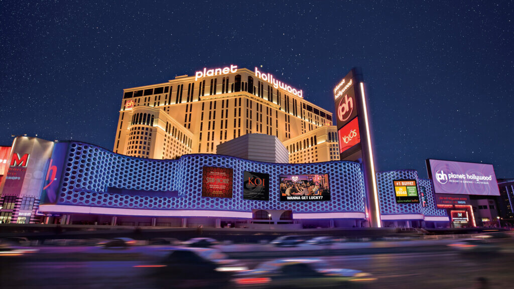 Night view of Planet Hollywood Resort & Casino in Las Vegas, with bright lights, colorful signage, and moving cars in the foreground beneath a starry sky.