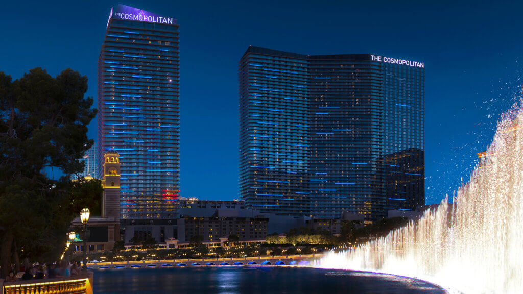 The Cosmopolitan hotel towers in Las Vegas are illuminated at night, with bright fountains from the Bellagio water show in the foreground against a dark blue sky.