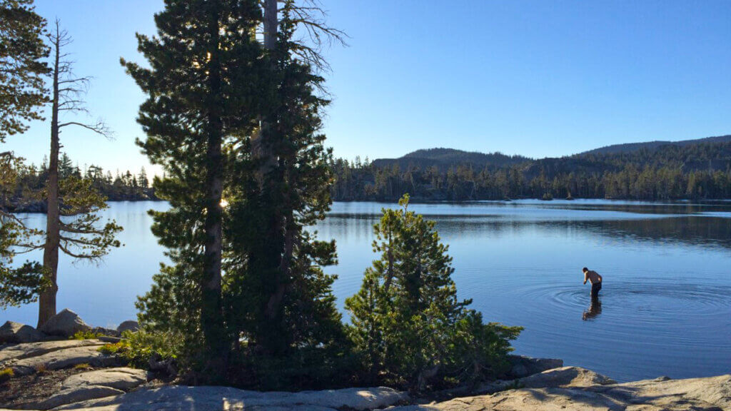 A person stands in shallow water near the shore of a calm lake surrounded by pine trees and hills under a clear blue sky.