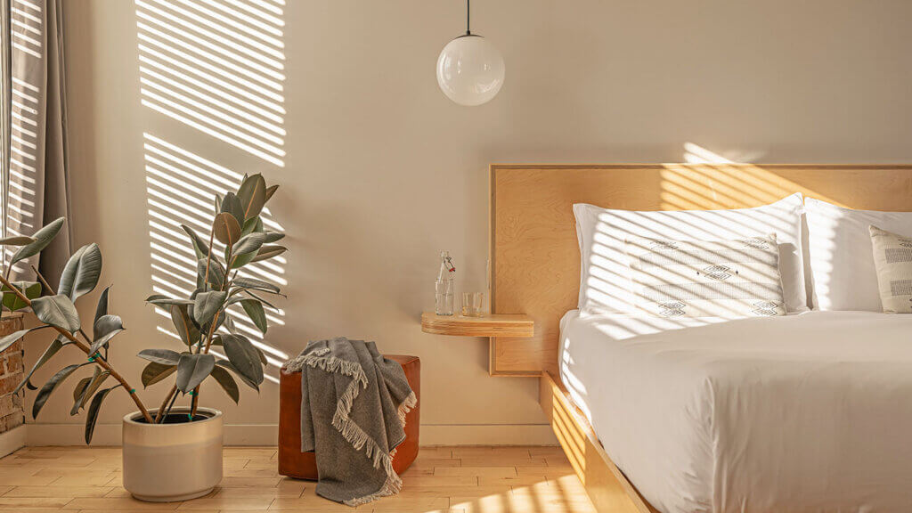 A sunlit minimalist bedroom with a white bed, wooden headboard, wall-mounted nightstand with water bottle and glasses, a potted plant, and a blanket draped over a brown pouf. Horizontal shadows from blinds cross the room.
