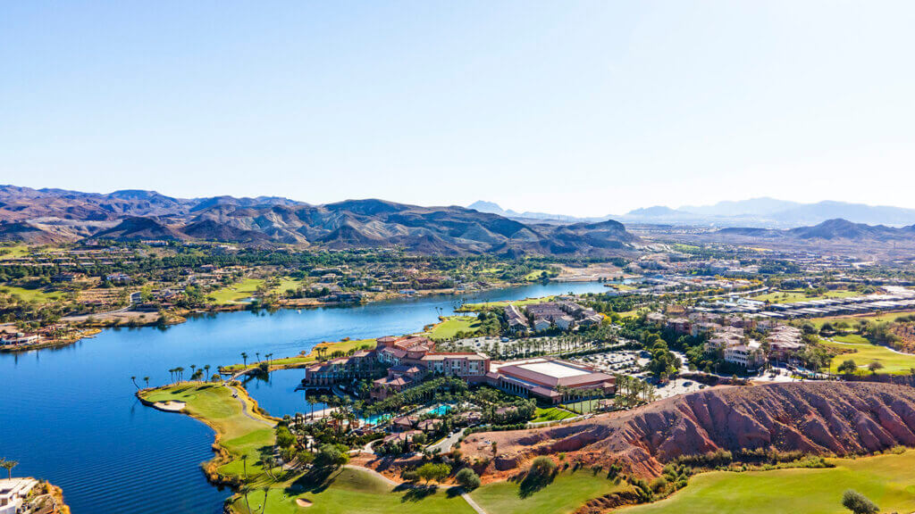 Aerial view of a scenic resort by a lake, surrounded by green golf courses, palm trees, and mountains in the distance under a clear blue sky.