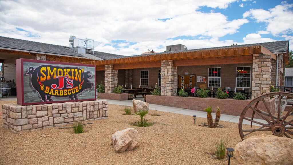 A stone sign for Smokin Js Barbeque stands in front of a rustic restaurant with a wooden pergola, stone pillars, and desert landscaping under a partly cloudy sky.