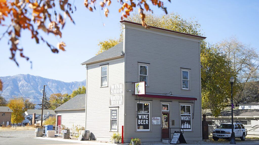 A two-story gray building with signs for wine, beer, and whisky stands on a sunny street with autumn leaves in the foreground and mountains in the background.