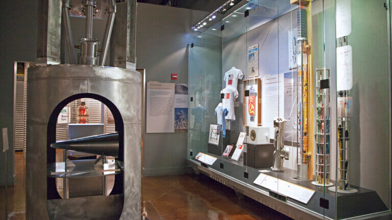 A museum exhibit displays various space exploration artifacts, including a metallic rocket nose cone, technical equipment, informational panels, and astronaut memorabilia in glass cases.