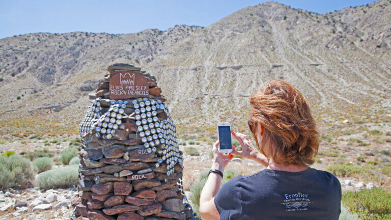 A woman takes a photo of a rock-covered monument with a sign reading Elvis Presley Rock n Roll in a desert landscape, with a mountain in the background.