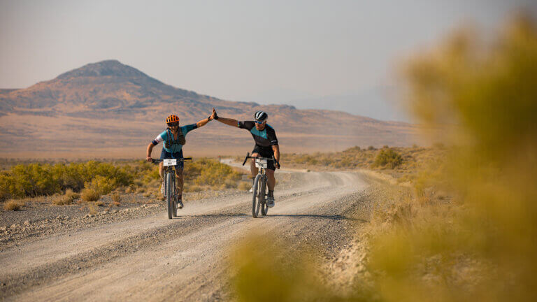 Two cyclists ride on a gravel road through a desert landscape, giving each other a high five, with distant mountains in the background and shrubs lining the road.