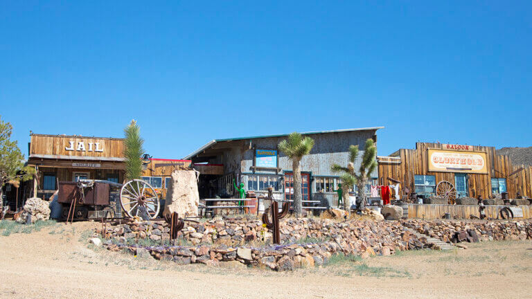 A row of old Western-style buildings, including a jail and saloon, stand behind a rocky, desert landscape with scattered cacti, wagon wheels, and sparse trees under a clear blue sky.