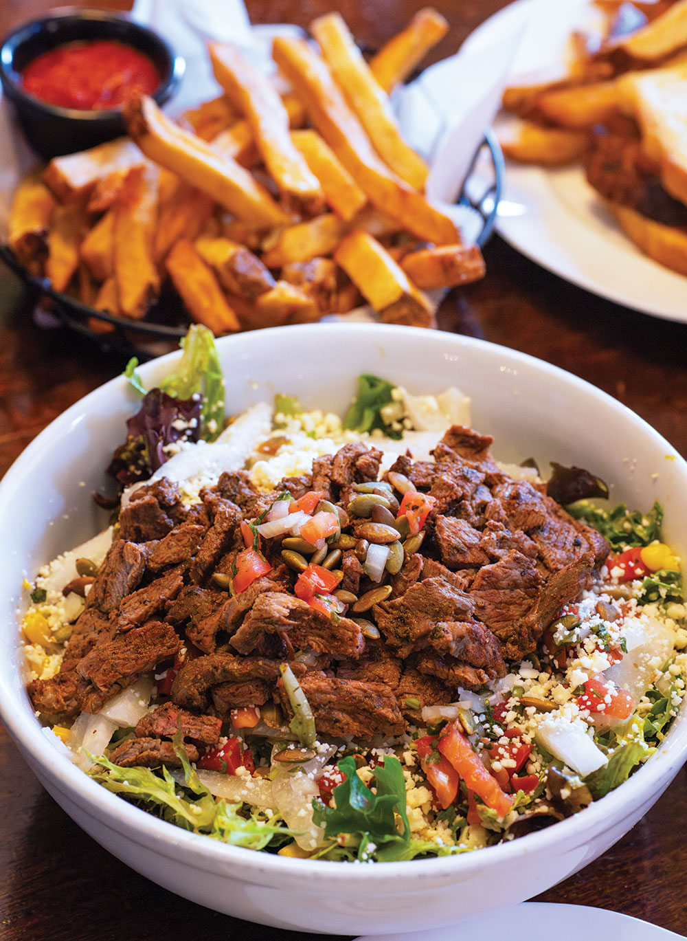 A bowl with beef and chopped salad, with french fries in the background