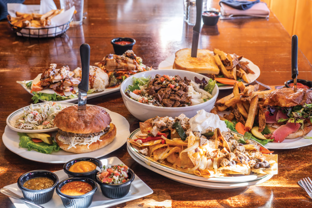 A wooden table full of food, served at Sassafras Eclectic Food Joint. Burgers, nachos, Asian food bowls, tacos, sandwiches, and various sauces.