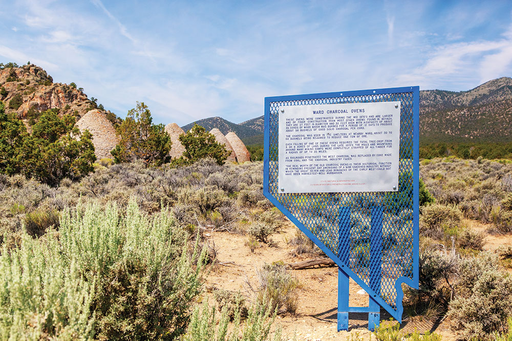 Blue metal state-of-Nevada sign with information about Ward Charcoal Ovens on it. The ovens are visible in the background.