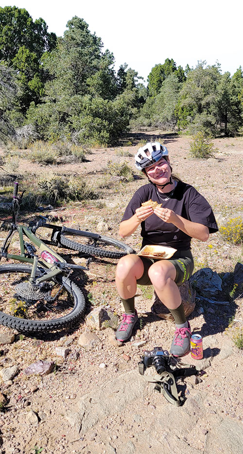A smiling mountain biker sits on a rock, eating lunch, with their bike laying beside them