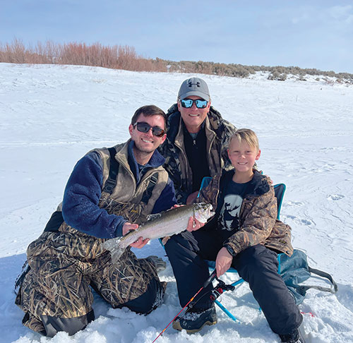 Two adults and a child - holding a fishing pole - smile for the camera, while holding a large fish. They sit/kneel in the snow, and there are desert willows behind them. Photo ©Nevada Department of Wildlife