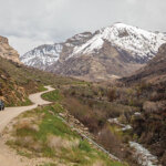 A car is on a mountain road that leads off into the distance, to a snowcapped peak. A river runs alongside the road.