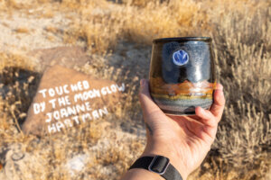 Hand holding a mug in front of a rock that says 'touched by the moonglow - Joanne Hartman.'