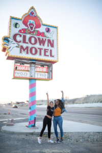 Two women standing next to the Clown Motel sign, pointing up to it.