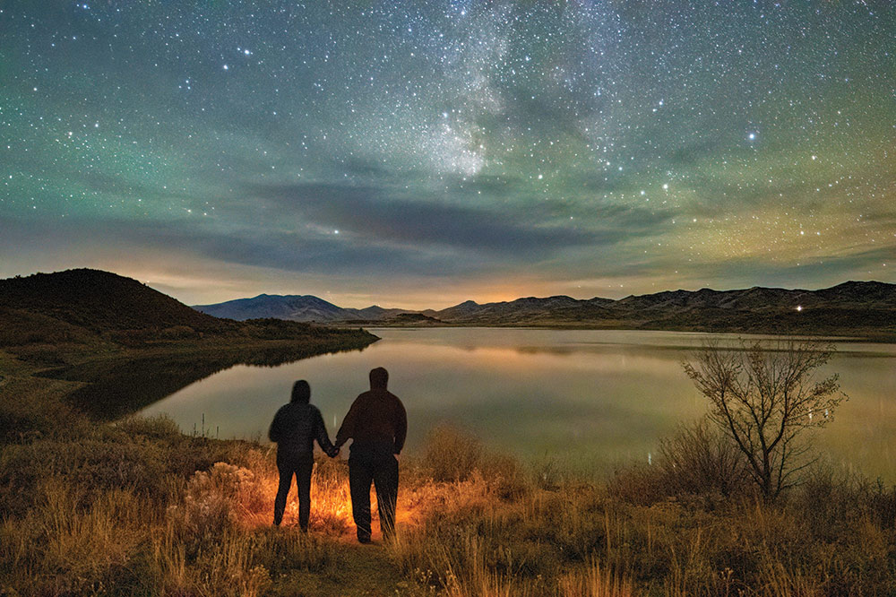 A couple is silhouetted on the shoreline of a calm Wild Horse Reservoir. Mountains surround and there are stars in the sky, including the Milky Way. Photo ©Jackie Gorton