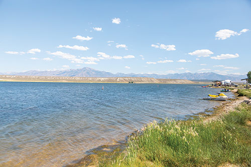 South Fork Reservoir shoreline with jet-skis, campers, kayaks, and lots of other recreational tools. There is tall green grass along the shore and mountains in the distance, with puffy white clouds in the sky.