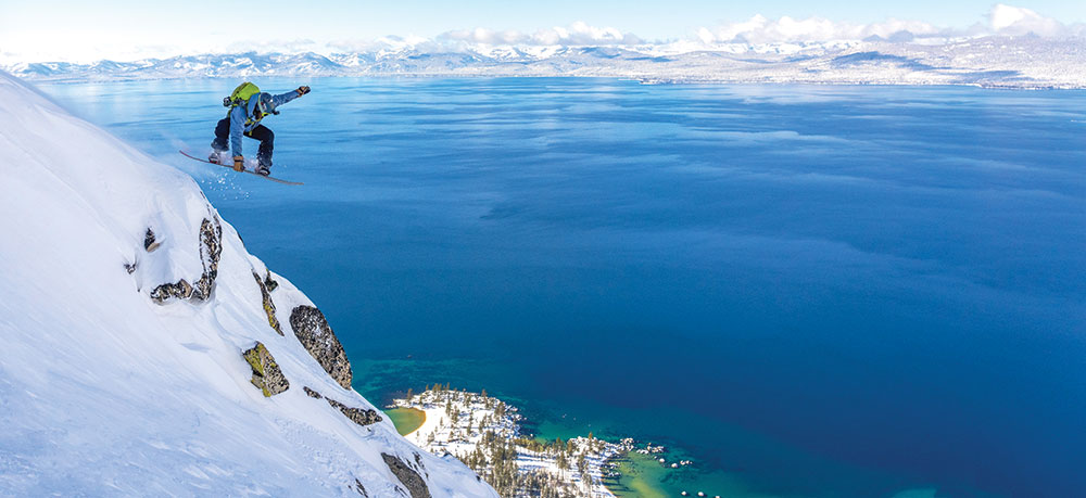 A snowboarder coming off a hill above Lake Tahoe, looking like he's jumping off into nothingness. Sand Harbor down below, with deep blue water and snowy mountains in the background.