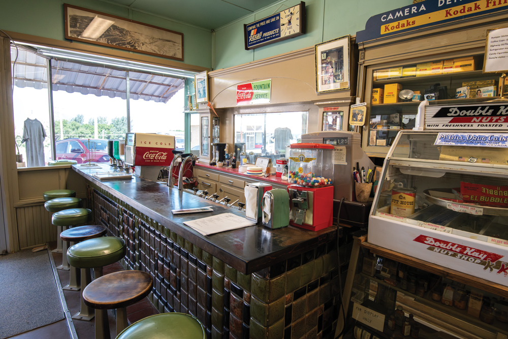 Inside McGill Drugstore Museum. Image shows a counter with 7 bar stools in front of said counter, with various decorations adorning the wall, while a soda machine, gumball machine, and napkin holder sits on top of the counter. Behind the counter is a cabinet full of film photography equipment alongside other random various items. A food display case sits beside the counter.