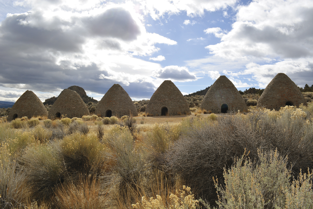 Stone beehive-shaped structures stand in a row under a cloudy sky, surrounded by desert vegetation.