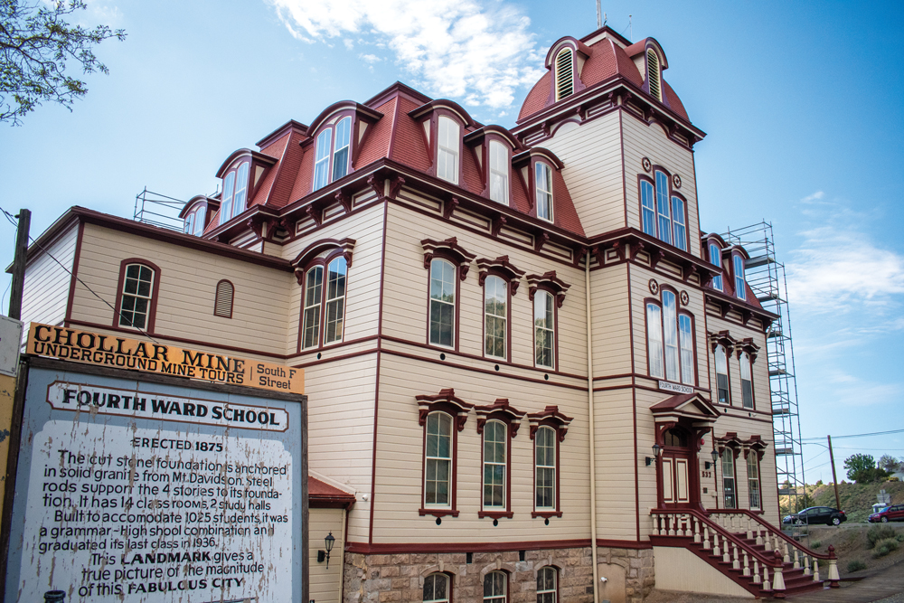 Exterior shot of Fourth Ward School. Taken at a 3/4th angle. The building has three floors including a basement. There is stairs that lead up to the main door, which has a decorative overhang roof. The main door area juts out a few feet from the rest of the building. Above the main door is three rectangular windows, and the windows repeat the same pattern on the floor above. On top of this section sits a decorative roof with two dormers extending off the roof. The roof is a red color while the reset of the structure is a beige. The rest of the walls that are set back from the main entrance have 3 windows each side and on the first and second floor. On the third floor, there are more dormers jutting out from the roof on each side. There is an extension to the left of the building with a flatter roof but only contains one window. There is a sign that is in front of the school that covering the rest of the extension. The sign has a narrow yellow sign on top of it that reads "Chollar Mine" in large letters with "Underground mine tours" just below it. to the right of these section says "South F Street". The main sign has a blue background and has a decorative border around the words "Fourth Ward School." Below this headline has information about the school. To the right of the building, there is scaffolding. The sky is blue with sparse white clouds. 