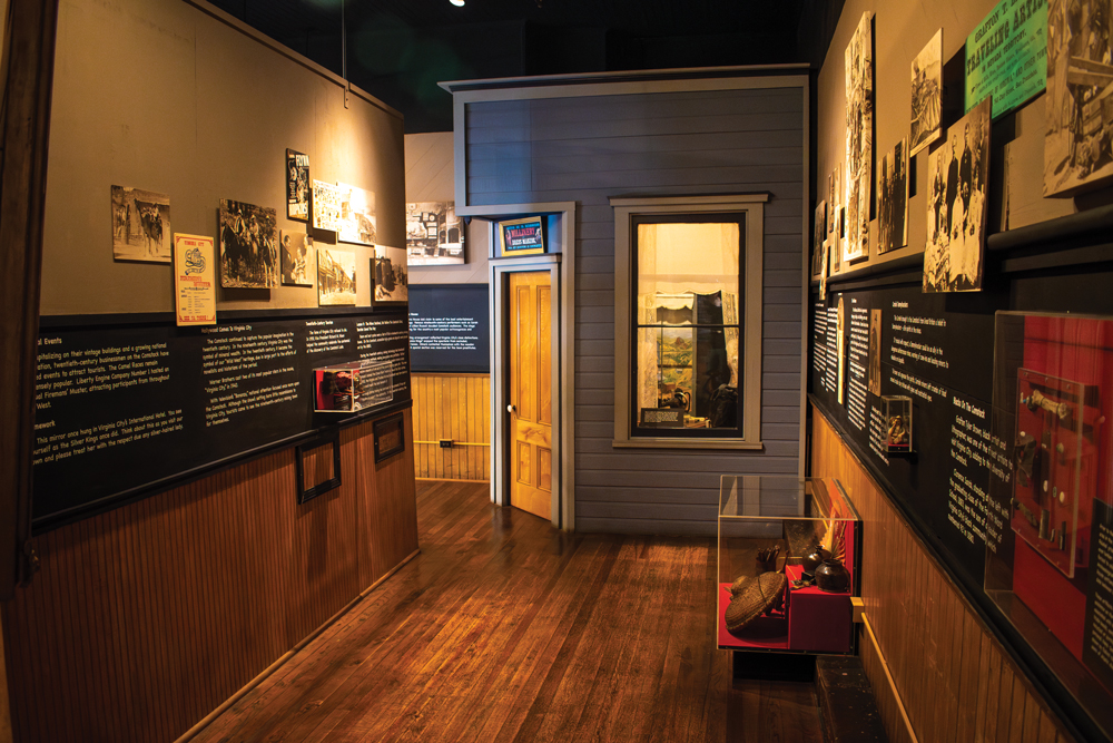 Museum Exhibit of Fourth Ward School. On the left hand side of the room, information about the school and pictures are displayed on the wall. Between the left hand wall and the back wall, there is a hallway that leads to another part of the the museum with more information, and a door sits on the corner of the back wall leading into another room with display cases. On the right hand wall, more pictures and information are displayed on the wall while a glass case sits pushed up to the wall, which features pottery. 