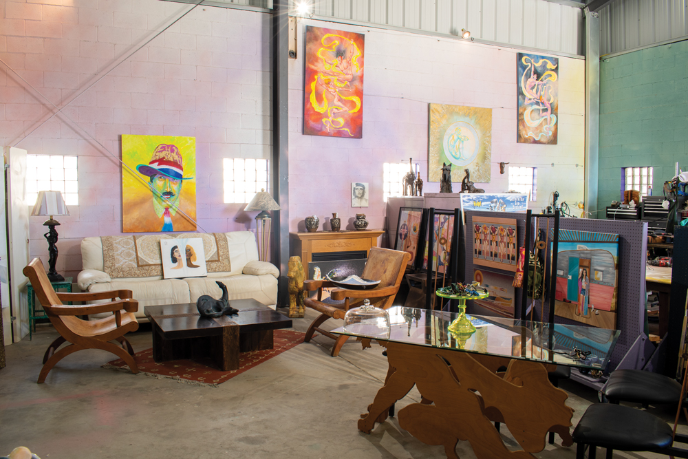 Interior of Bizarre 101 in Walker. The scene features an eclectic array of furniture. A white couch with a floral patterned fabric sits along the back wall of the interior. A painting that has two halves of heads sit on top of the couch. Above the couch is a painting featuring a man in a fedora. To the left of the couch, there is a green night stand type table with a large lamp sitting on top of it. In front of the couch, there are two wooden chairs and a wooden coffee table with a statue on top of it. There is a red rug underneath the coffee table. To the right of the couch sits another lamp and a wooden fireplace with three vases on top of it. Above the fireplace are two paintings; one is a painting of a man, the other is a painting of a female dancer with yellow ribbons flowing around her. To the right of the fireplace is a wall divider that has five paintings attached to it. The four paintings closest to the fireplace are abstract pieces. The last one is a painting of a retro style camper painted pink on the bottom with a silver top, a blue door attached to the camper, and a woman with black hair standing in front of the camper. Next to the paintings sits a table with an abstract base and a glass top. On top of the table is a green cake stand with knick knacks resting on top of it. There are two dinner table style chairs with black leather seats sitting to the right of the table. Towards the back of the photo sit two more large paintings, one is gold and has a white orb on it, while the other is another woman dancing with yellow ribbons that turn blue around her. The walls are painted in a gradient style fashion with pink, lavendar, and yellow on one wall; green on the other. There are 5 windows which allow light in.