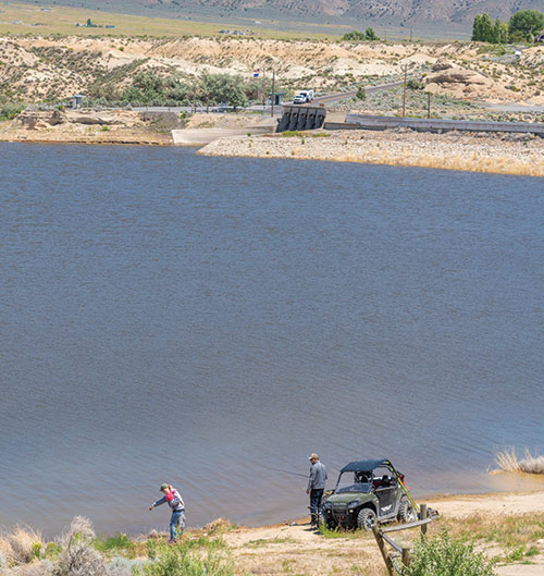 People fishing along the shore of Rye Patch Reservoir. There is a UTV parked nearby, and one person holds a child, while pointing into the water.