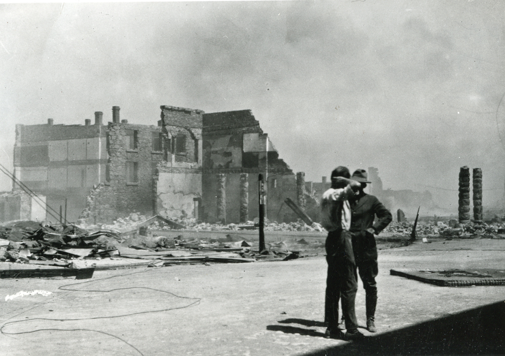 An old photo of two firemen standing on a street in front of a half ruined building with smoke rising from it. Photo was taken in 1923 Goldfield fire incident.