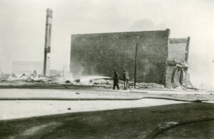 Two men can be seen with a firehose in their hands trying to put out a fire. Behind them stands a ruined building with brick walls still standing while everything else has been gutted on the building.