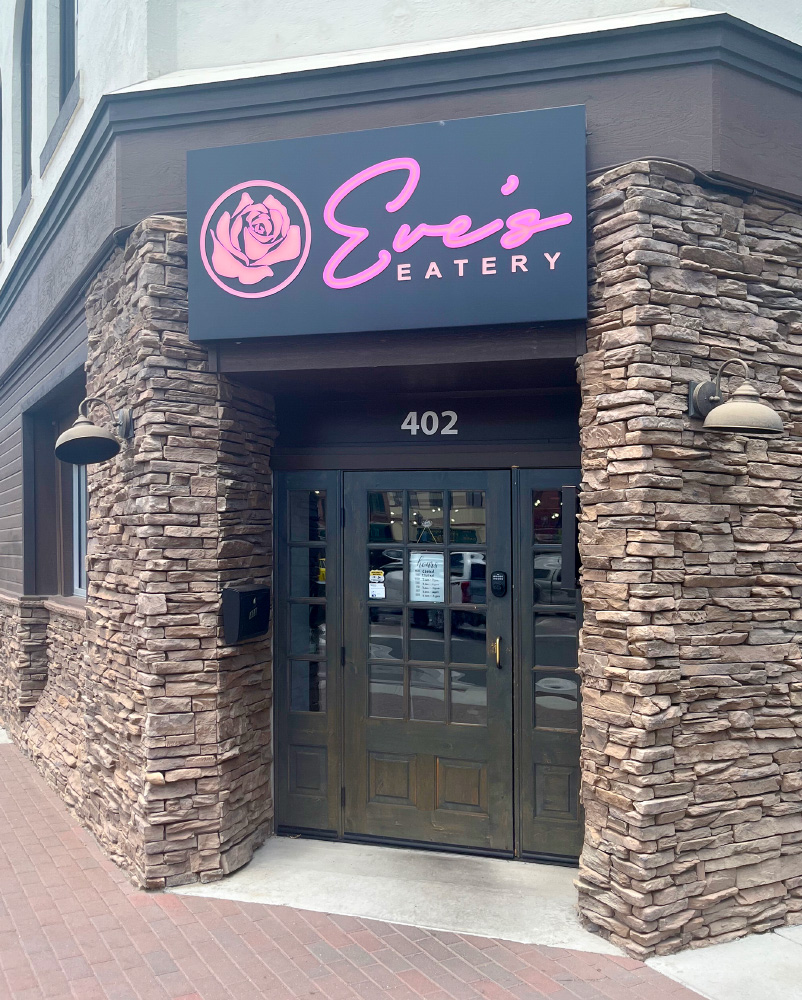 Historic exterior of Eve's Eatery. Tan stone walls with pink and black sign above the door.