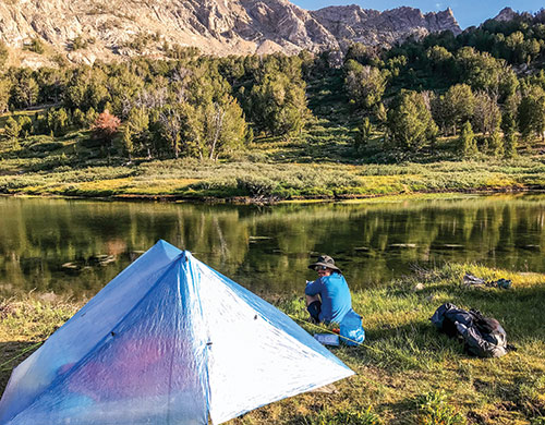 A young man, turned around to face the camera, sits in front of a tent on the edge of an alpine lake with a lot of trees and tall mountains surrounding.