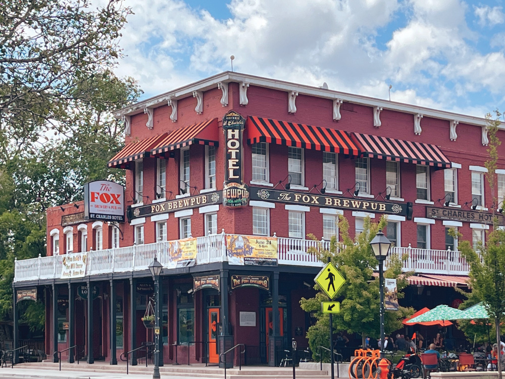 Exterior photo of The Fox Brewery & Pub. Red brick historic building, with puffy clouds in the blue sky, trees all around, and people sitting out under umbrellas, eating.