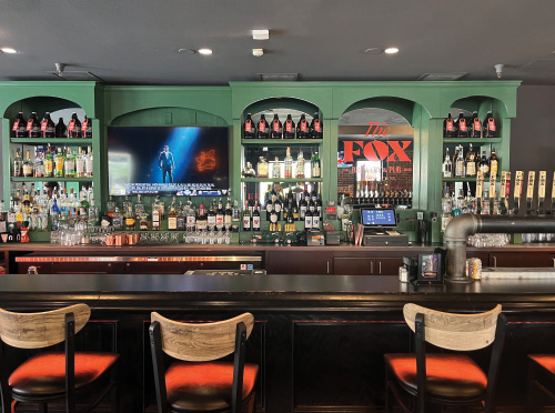 An interior photo of the Fox Brewery & Pub. Wooden bar top with stools and green bar with various bottles of alcohol.