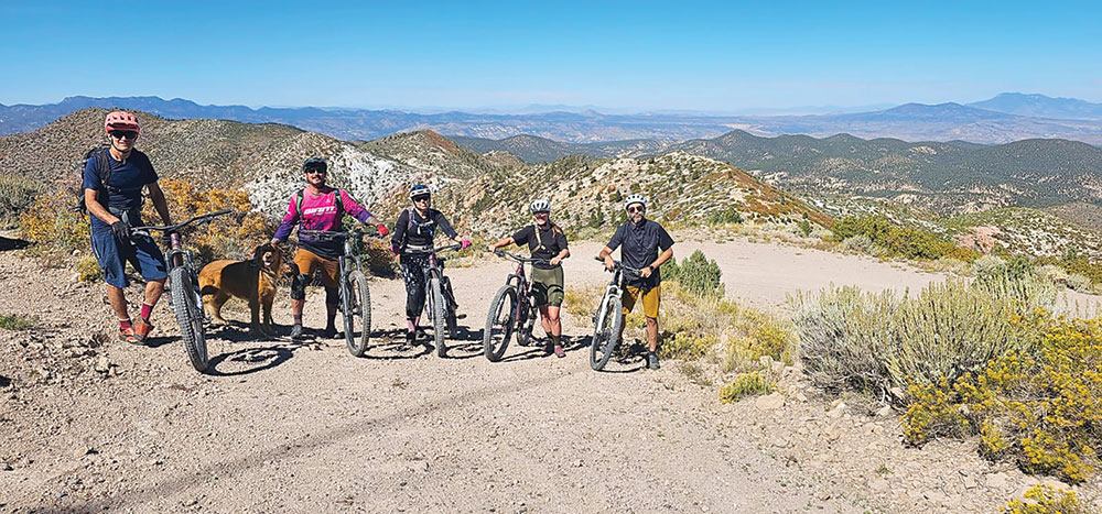 A group of mountain bikers and a dog, posing on a dirt road on Ella Mountain, with layers of mountains in the distance.