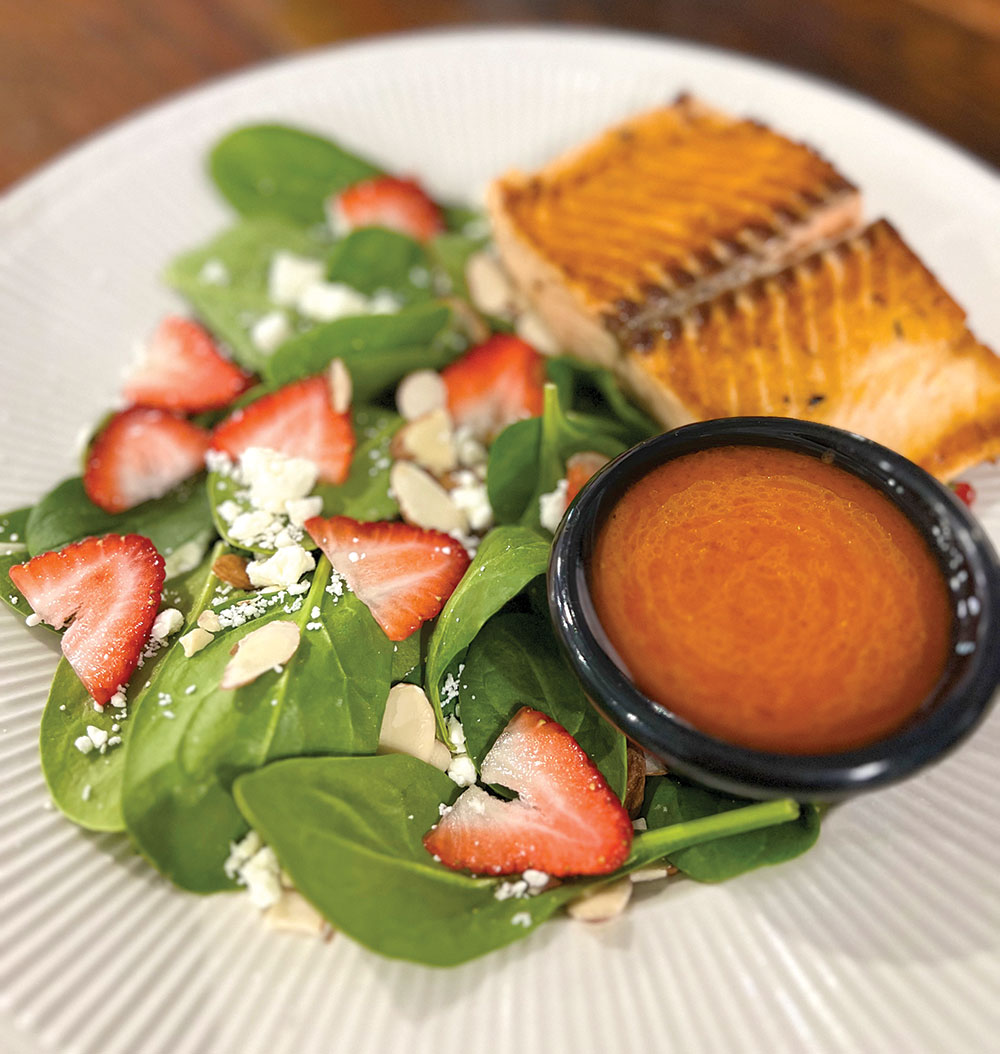 A piece of fish with a spinach salad that has sliced strawberries, crumbled cheese and blood orange vinaigrette dressing on the side