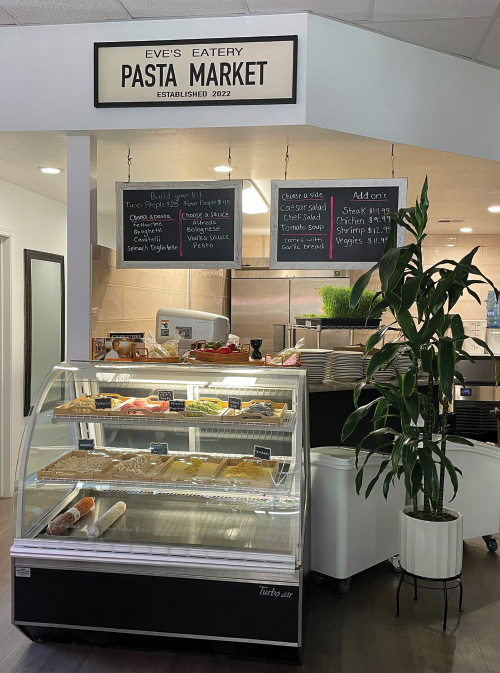 Interior of Eve's Eatery. Display case with several types of homemade pasta, with a sign that reads "Pasta Market" above it. Plants and plates also visible.